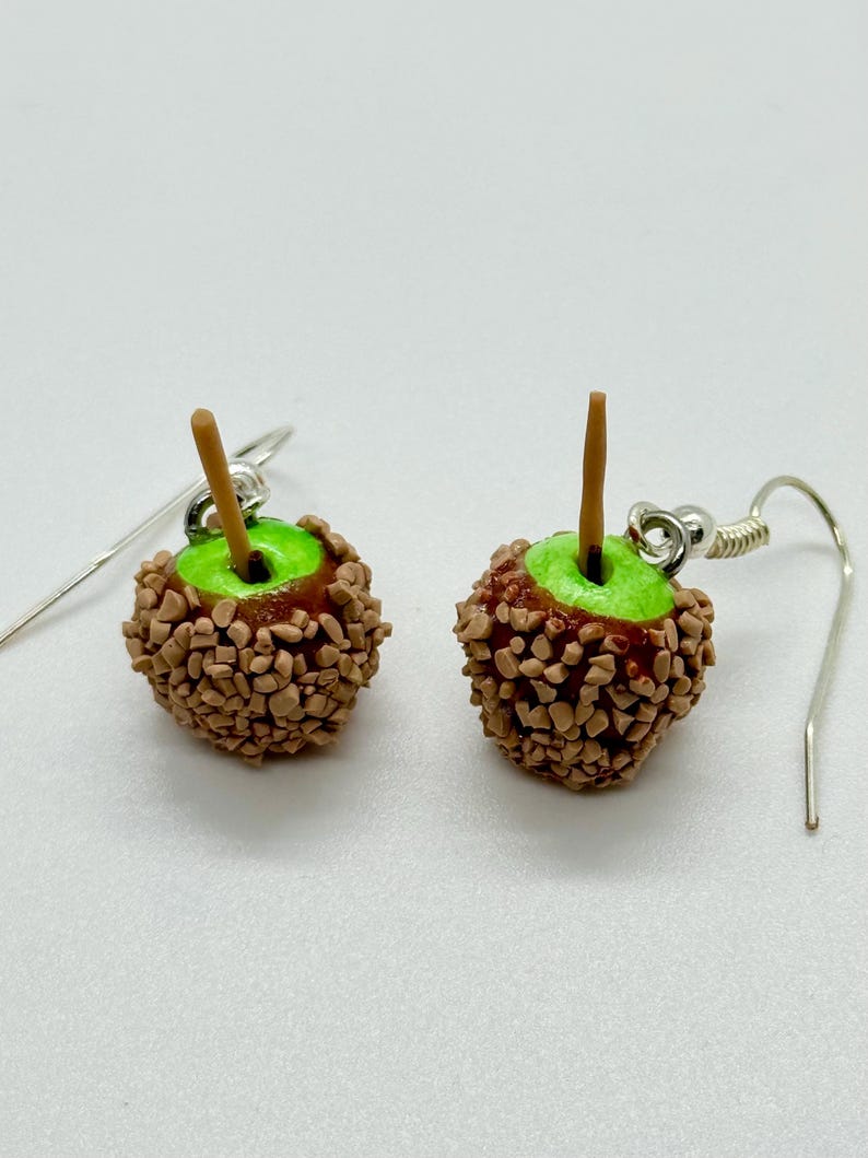May include: A pair of handmade caramel apple earrings. Each earring features a miniature green apple base, coated in a brown candy shell with a wooden stick. The earrings have silver-colored ear wires.