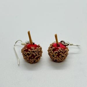 May include: A pair of dangle earrings shaped like miniature caramel apples. Each earring features a red apple base covered in brown candy pieces, with a small brown stick. The earrings hang from silver-colored fish hook ear wires.