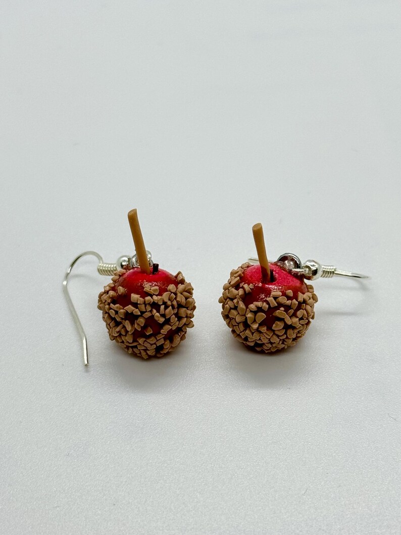 May include: A pair of dangle earrings shaped like miniature caramel apples. Each earring features a red apple base covered in brown candy pieces, with a small brown stick. The earrings hang from silver-colored fish hook ear wires.