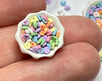 Può includere: Una piccola ciotola bianca piena di cuori di caramelle colorati. I cuori di caramelle sono in varie tonalità di rosa, rosso, verde, giallo e blu. La ciotola è appoggiata su un dito.