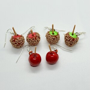 May include: A collection of handmade apple-shaped earrings. Some are glossy red with a wooden stick, others are covered in brown sprinkles with a red center, and some have green centers. The earrings have silver hooks.