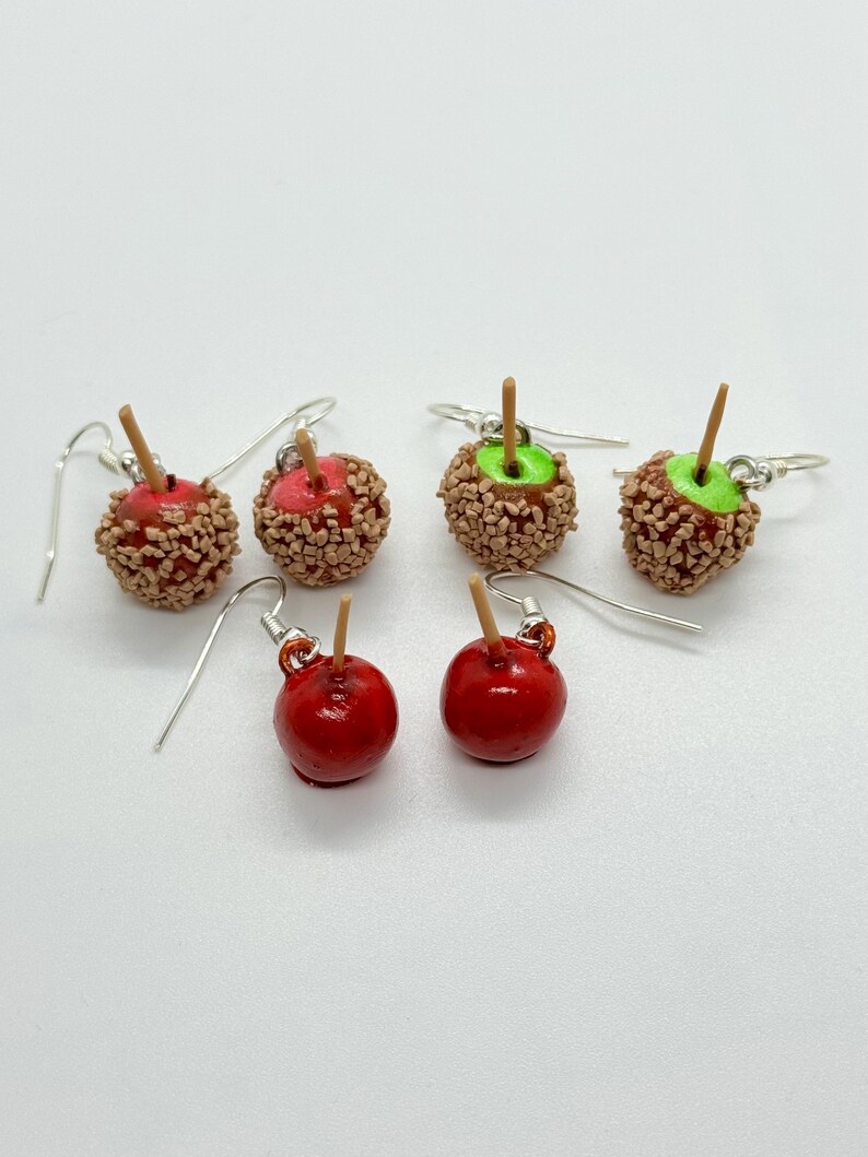 May include: A collection of handmade apple-shaped earrings. Some are glossy red with a wooden stick, others are covered in brown sprinkles with a red center, and some have green centers. The earrings have silver hooks.