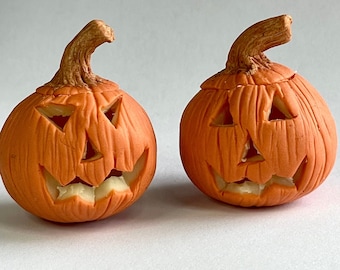 Puede incluir: Dos figuritas de cerámica naranja en forma de calabaza de Halloween con caras talladas. Las figuritas están sobre un fondo blanco.