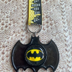 Puede incluir: Un llavero con el logotipo de Batman negro con un cordón de paisaje urbano amarillo y negro. El logotipo de Batman es amarillo sobre un fondo negro, dentro de un marco negro en forma de murciélago. El cordón es amarillo con un patrón de paisaje urbano negro.