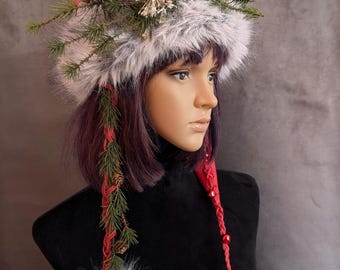May include: A winter-themed headpiece with a faux fur base, adorned with pine needles, small pine cones, and red accents. The headpiece has two dangling faux fur pom-poms. The Etsy shop name "originalsbyeva.etsy.com" is visible.