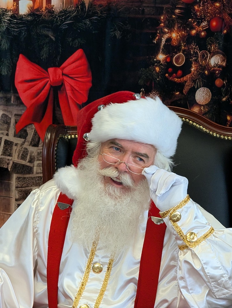 May include: A Santa Claus wearing a white suit with red suspenders and a white fur-trimmed hat. He is sitting in a chair and has a white beard and mustache. He is wearing glasses and looking at the camera.