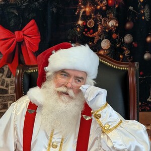 May include: Santa Claus wearing a white suit with red suspenders and a white beard. He is wearing a red and white hat with bells and is holding his glasses with his right hand. He is sitting in a black leather chair in front of a Christmas tree.