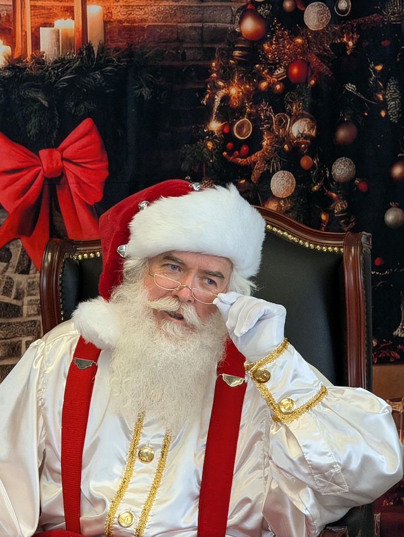 May include: Santa Claus wearing a white suit with red suspenders and a white beard. He is wearing a red and white hat with bells and is holding his glasses with his right hand. He is sitting in a black leather chair in front of a Christmas tree.