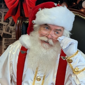 May include: A Santa Claus wearing a white fur-trimmed hat, red suit, and white gloves. He is looking at the camera and holding his glasses with his right hand. He has a white beard and mustache.
