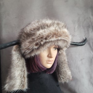 May include: A gray and brown faux fur hat with ear flaps and a black leather headband. The hat has a unique design with the headband extending out on either side to form horns.