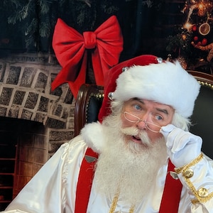 May include: Santa Claus wearing a white suit with red suspenders and a white beard. He is wearing a red hat with white trim and silver bells. He is holding his glasses with his right hand and looking to the left.
