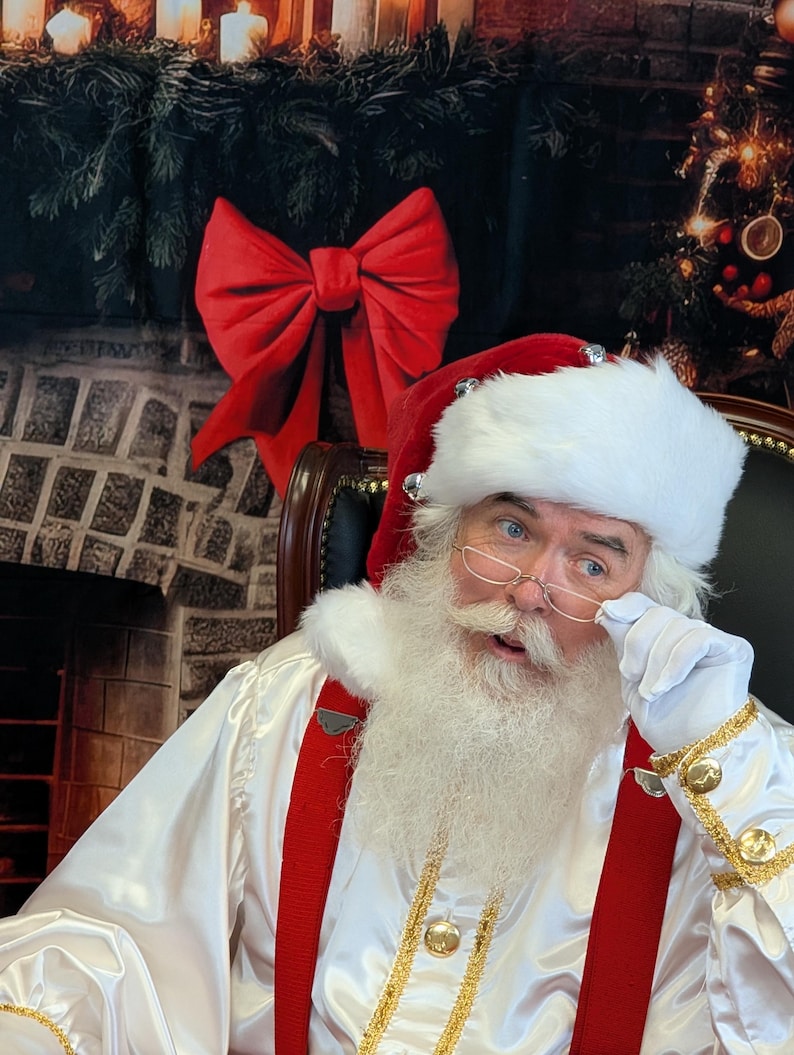 May include: Santa Claus wearing a white suit with red suspenders and a white beard. He is wearing a red hat with white trim and silver bells. He is holding his glasses with his right hand and looking to the left.