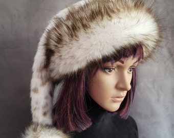 May include: A white and brown faux fur hat with a long, fluffy tail and a pom-pom on the end. The hat is worn on a mannequin with purple hair.