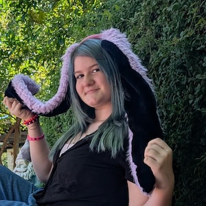 May include: A plush bunny ear hat with long, floppy ears. The hat features black and lavender faux fur, with a pink interior. The person in the image is wearing the hat and smiling, with a green background.
