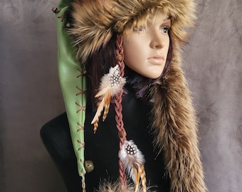 May include: A green faux leather hat with a brown faux fur trim and a long brown faux fur tail. The hat has a brown leather strap with a bell and feathers attached. The hat is on a mannequin head.
