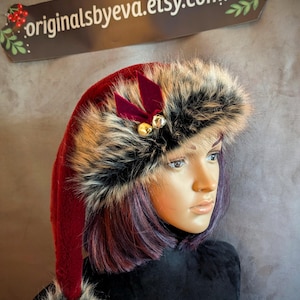 May include: A red Santa hat with a faux fur trim and a matching pom-pom. The hat is adorned with a dark red velvet bow and two small gold bells. The text "originalsbyeva.etsy.com" is on a brown tag.