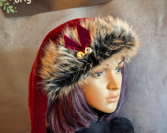 Può includere: Un cappello da Babbo Natale rosso con finiture in ecopelliccia e pompon abbinato. Il cappello è adornato con un fiocco di velluto rosso scuro e due piccole campane dorate. Il testo "originalsbyeva.etsy.com" è su un'etichetta marrone.