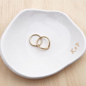 May include: A white ceramic ring dish with two gold rings. The dish has an irregular shape and features the gold initials "K+P". The rings are interlocked. The dish is likely for storing jewellery.