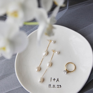 May include: A white ceramic ring dish with a gold ring and pearl earrings. The dish has the text "I + A 11.11.23" written on it.