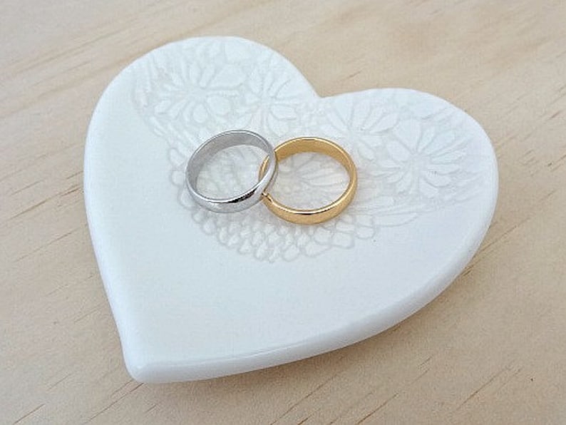 K&ouml;nnte beinhalten: Eine wei&szlig;e, herzf&ouml;rmige Ringhalterung aus Keramik mit floralem Design. Zwei Eheringe, einer silber und einer gold, liegen auf der Schale.