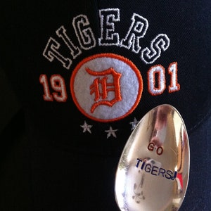 May include: A black baseball cap with white embroidered text that reads "TIGERS 1901" and a white and orange embroidered "D" inside a circle. A silver spoon with blue text that reads "GO TIGERS!" is in the foreground.