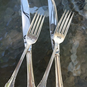 May include: Two silver forks with the words "MR" and "MRS" engraved on the handles, and a silver knife. The forks and knife are arranged in an "X" shape on a textured surface.