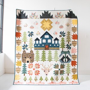 Peut inclure: Une courtepointe faite à la main sur le thème de la forêt. La courtepointe présente une maison bleue, des arbres, un ours, un raton laveur et un cerf. La courtepointe a un fond blanc avec des accents colorés d'orange, de jaune, de vert et de marron. La courtepointe est accrochée à un mur blanc.