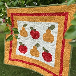 May include: A quilted wall hanging featuring a repeating pattern of apples and pears. The quilt has a white center with red and yellow fruit designs. The border is yellow with white polka dots, and a red inner border.