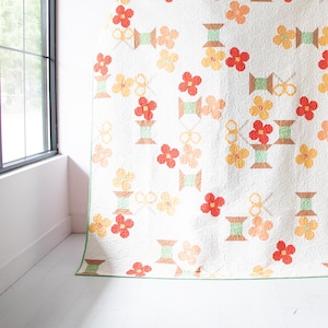 May include: A white quilt with a repeating pattern of red, orange, and yellow flowers and green spools of thread. The quilt is hanging on a white wall.