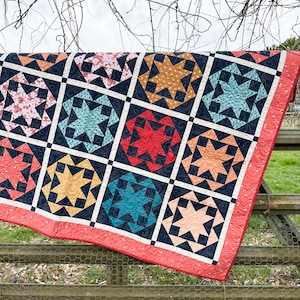 May include: A colourful quilt with a geometric pattern of stars and squares. The quilt is made with a variety of fabrics in shades of red, orange, yellow, blue, green, and black. The quilt is hanging on a wooden fence.