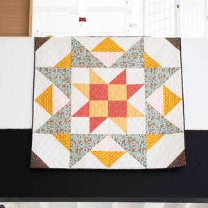 May include: A white quilt with a large star in the center. The star is made of red and yellow squares. The quilt is surrounded by a border of brown fabric.