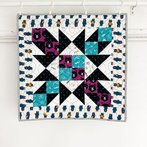 May include: A square quilt with a starburst design. The centre features black triangles and squares of teal and purple fabric with bear patterns. The border has a repeating bear pattern on a white background, with a grey striped edge.