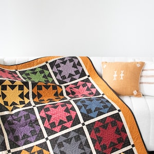 May include: A colourful quilt with a starburst pattern in shades of red, orange, yellow, green, blue, and purple. The quilt is folded and draped over a white sofa with cushions.