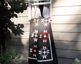 May include: A black and white quilt with pink, red, and yellow star-shaped patterns hanging on a light brown wooden wall.