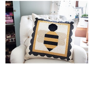 Puede incluir: Una almohada de abeja negra y amarilla con un borde festoneado. La almohada mide 61 cm por 61 cm y está hecha de tela con un patrón de lunares negros y blancos. La almohada está sentada en un sillón blanco. El texto "Just Buzzin' Around" y "24" x 24" Pillow -or- Quilt" está por encima de la almohada. El texto "Lamb Farm Designs" y "By A.K. & J.J." está debajo de la almohada.