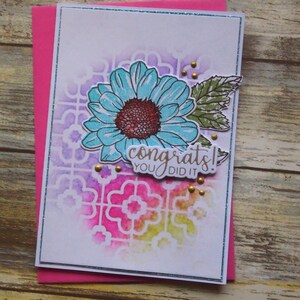 May include: A handmade greeting card featuring a blue flower and the words "congrats! you did it." The card has a white background with a colourful pattern and is set on a pink envelope. Gold embellishments add a touch of elegance.