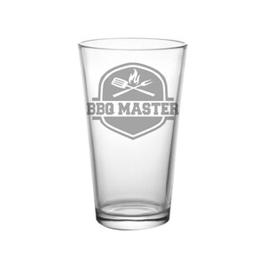 May include: A clear pint glass with a gray BBQ Master logo. The logo features a shield shape with a spatula, fork, and flames. The glass is empty and stands upright against a white background.