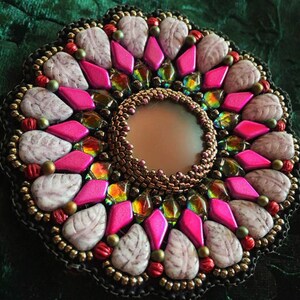 Pode incluir: Um broche circular de contas com um esquema de cores rosa, branco e dourado. O broche apresenta um espelho redondo central rodeado por um padrão geométrico de contas e pedras.