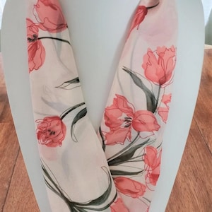 May include: A white silk scarf with a floral print of pink tulips. The scarf is folded in a V-shape and displayed on a white mannequin.