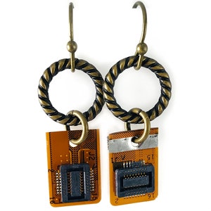 Puede incluir: Un par de pendientes de color dorado con un diseño de cuerda trenzada. Cada pendiente tiene un pequeño circuito impreso con un chip negro y el texto "J2" y "J1".