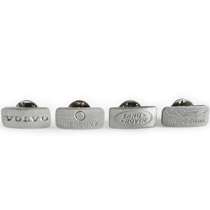 Pode incluir: Quatro broches de lapela prateados com diferentes logotipos de carros. Os logotipos são Volvo, Mercury, Land Rover e Aston Martin.
