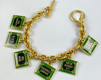 May include: Gold-toned charm bracelet featuring rectangular charms with black text on a green background. The charms display street names like "5th Ave." and "Rodeo Drive." The bracelet has a toggle clasp.