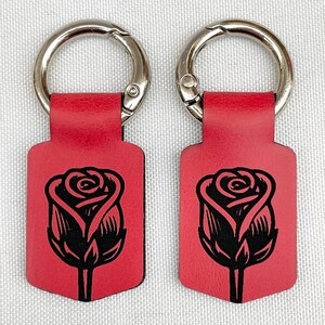May include: Two red leather keychains with a black rose design. Each keychain has a silver-colored metal clasp. The rose is a detailed illustration. The keychains are rectangular with a rounded bottom.