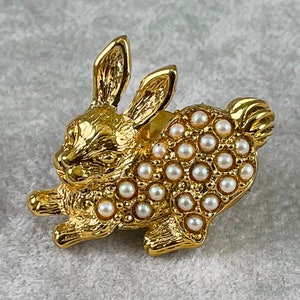 May include: A gold-toned rabbit brooch with a pearl-like finish. The rabbit is in a running pose with its ears up. The brooch is covered in small, round, white pearls.