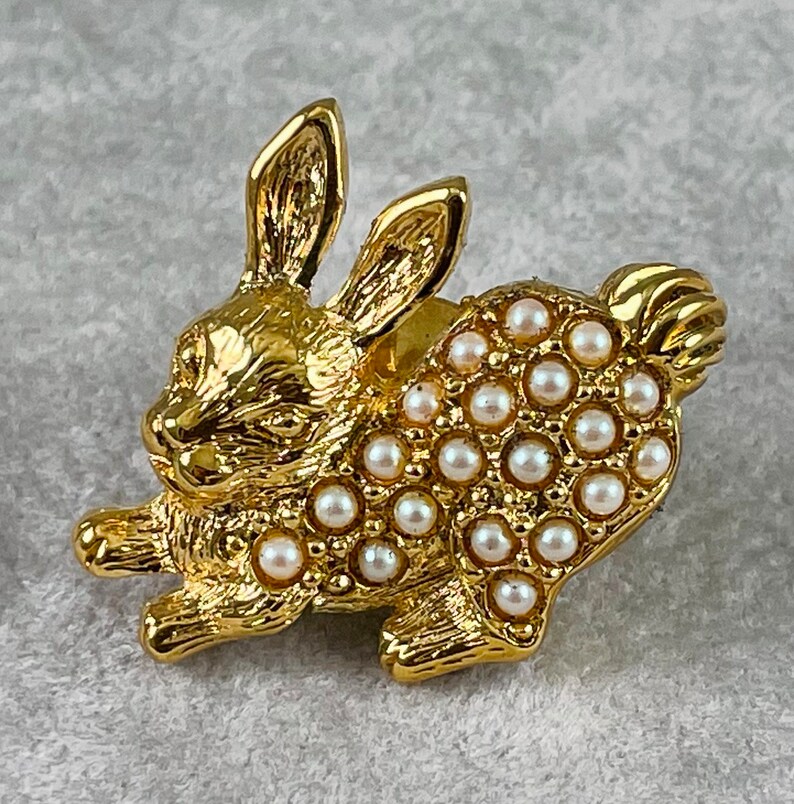 May include: A gold-toned rabbit brooch with a pearl-like finish. The rabbit is in a running pose with its ears up. The brooch is covered in small, round, white pearls.