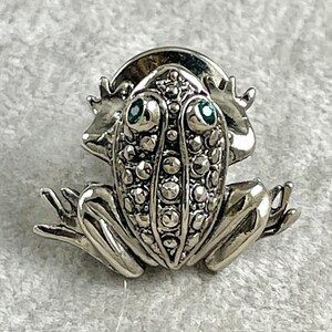 May include: A silver frog brooch with green gem eyes. The frog is detailed with a textured surface and has a rounded back.