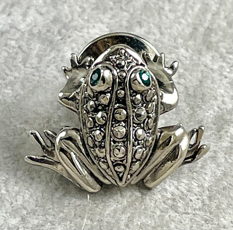 May include: A silver frog brooch with green gem eyes. The frog is detailed with a textured surface and has a rounded back.