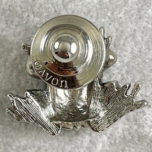 May include: Silver frog figurine wearing a hat, sitting on a white surface. The frog is wearing a hat with a round top and a wide brim. The frog is sitting on its hind legs, with its front legs outstretched. The frog's eyes are closed. The frog is made of metal and has a shiny finish. The frog is about 5 cm tall. The hat is about 2.5 cm in diameter. The frog is marked "AVON" on the bottom of the hat.
