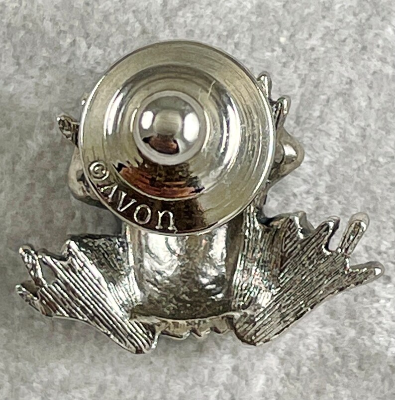 May include: Silver frog figurine wearing a hat, sitting on a white surface. The frog is wearing a hat with a round top and a wide brim. The frog is sitting on its hind legs, with its front legs outstretched. The frog's eyes are closed. The frog is made of metal and has a shiny finish. The frog is about 5 cm tall. The hat is about 2.5 cm in diameter. The frog is marked "AVON" on the bottom of the hat.