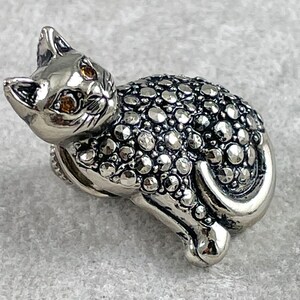 May include: A silver cat figurine with a detailed, textured surface covered in small, round stones. The cat has yellow eyes and a curled tail.